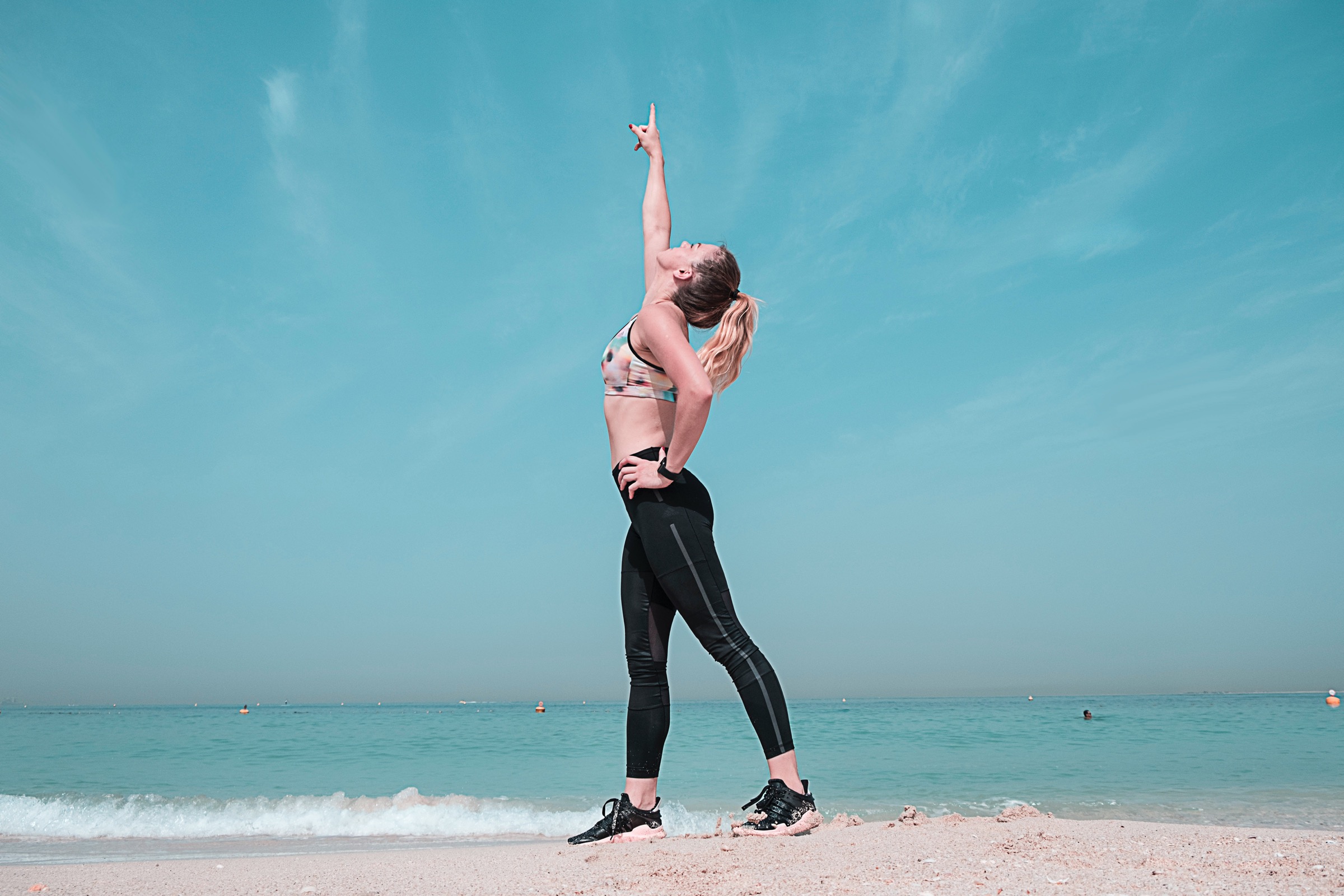 Woman wearing workout clothes at the beach while pointing up the sky 